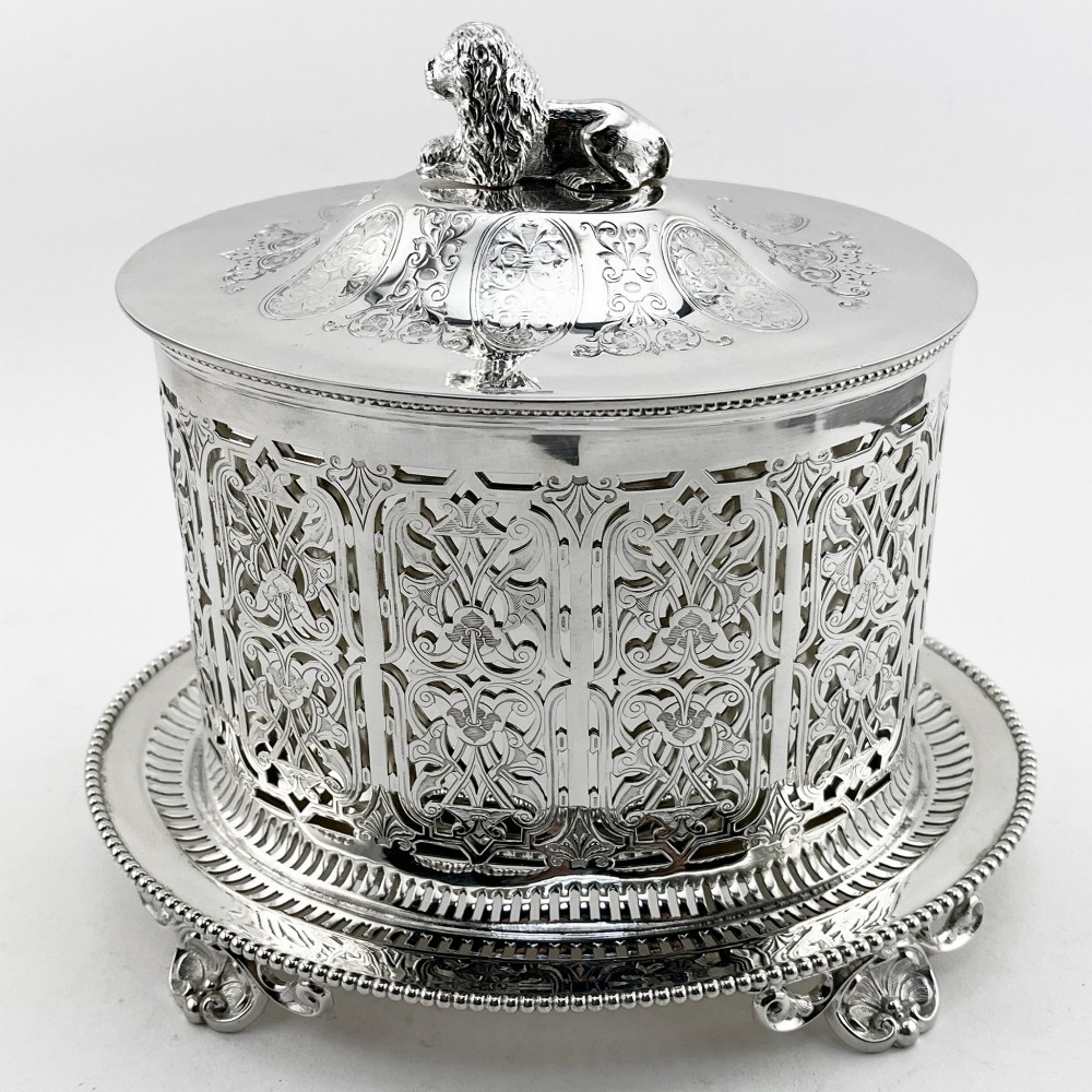 An oval Victorian silver-plated biscuit box featuring reticulated fretwork sides, an engraved lid with a dog finial, and a beaded footed base.