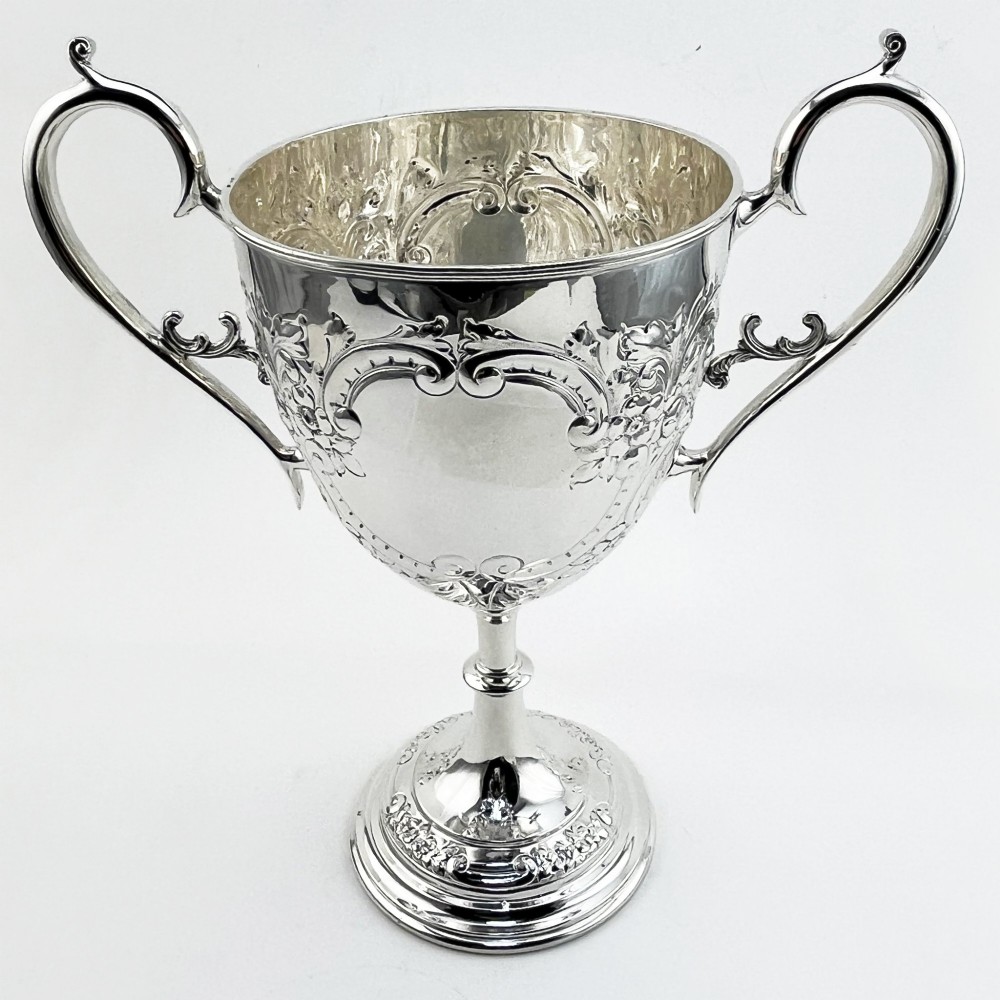 A silver-plated Victorian trophy cup featuring chased scrollwork, twin high-arched loop handles, and a stepped circular pedestal base.