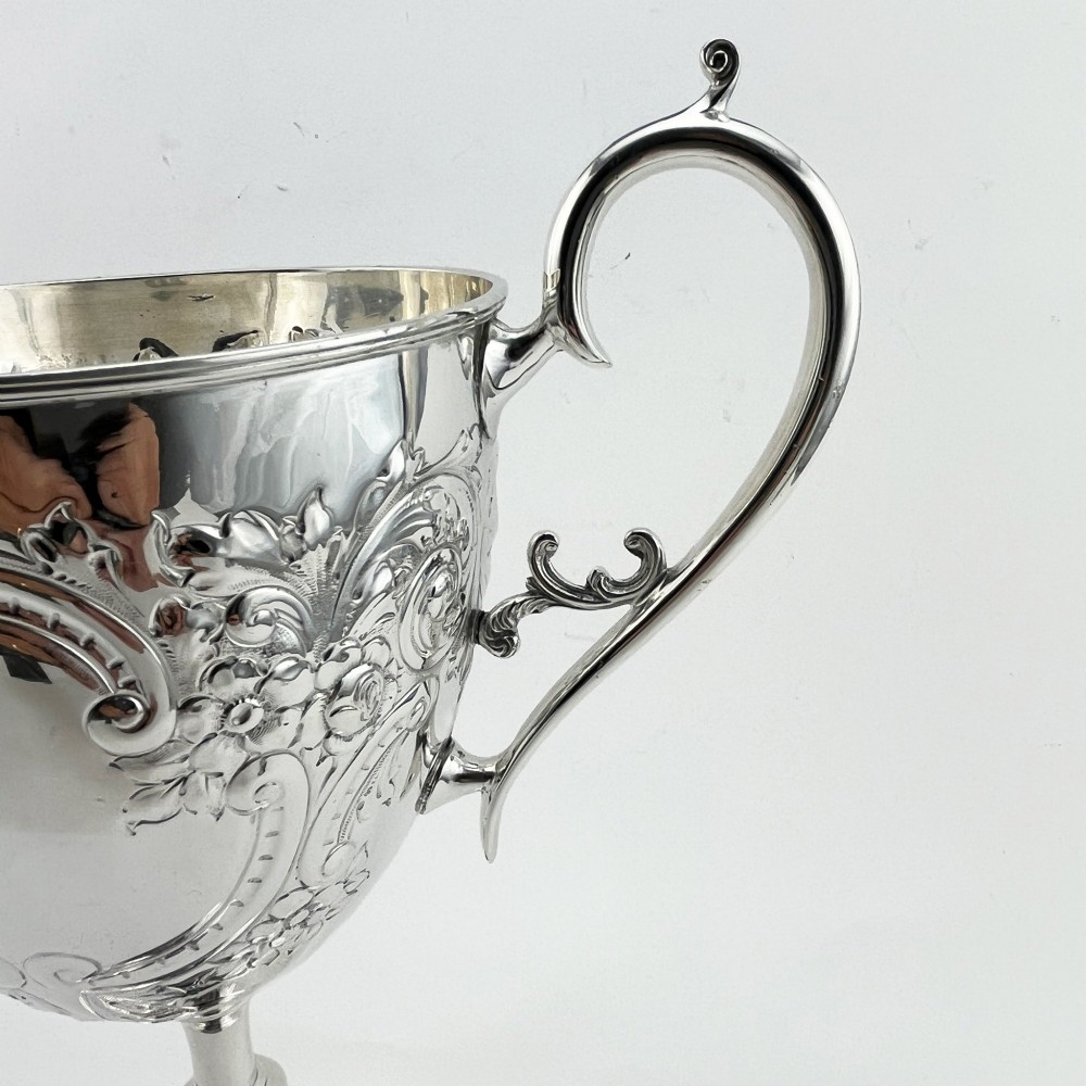 A silver-plated Victorian trophy cup featuring chased scrollwork, twin high-arched loop handles, and a stepped circular pedestal base. View II