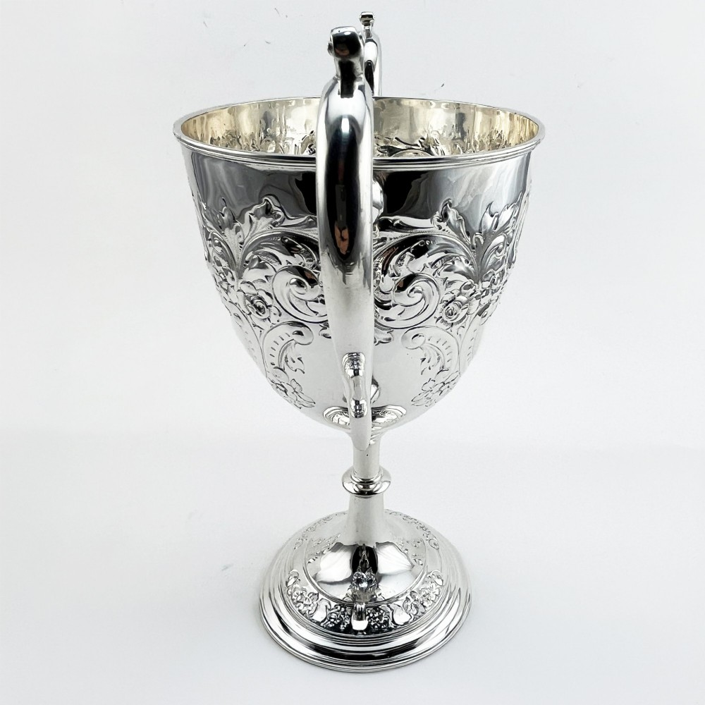 A silver-plated Victorian trophy cup featuring chased scrollwork, twin high-arched loop handles, and a stepped circular pedestal base. View III