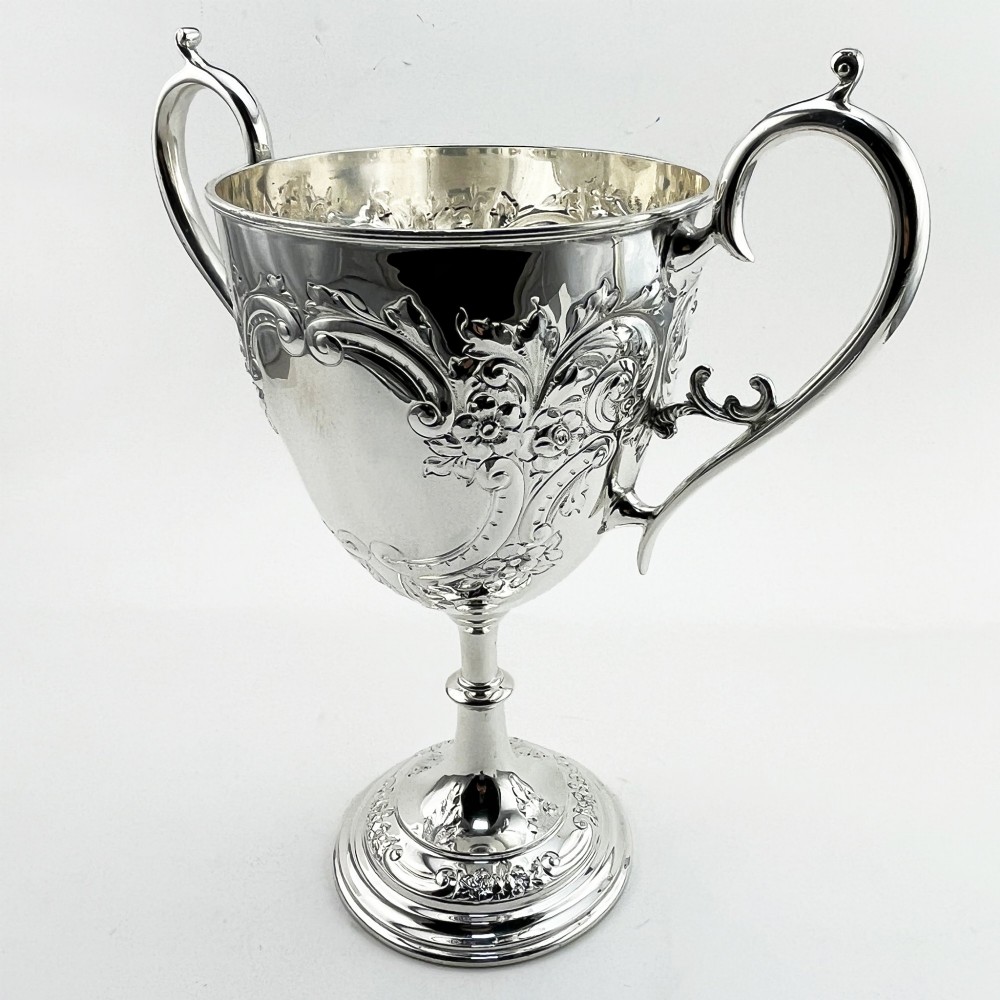 A silver-plated Victorian trophy cup featuring chased scrollwork, twin high-arched loop handles, and a stepped circular pedestal base. View IV