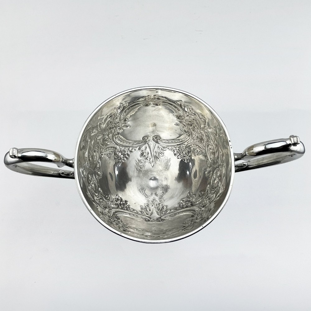 A silver-plated Victorian trophy cup featuring chased scrollwork, twin high-arched loop handles, and a stepped circular pedestal base. View VI