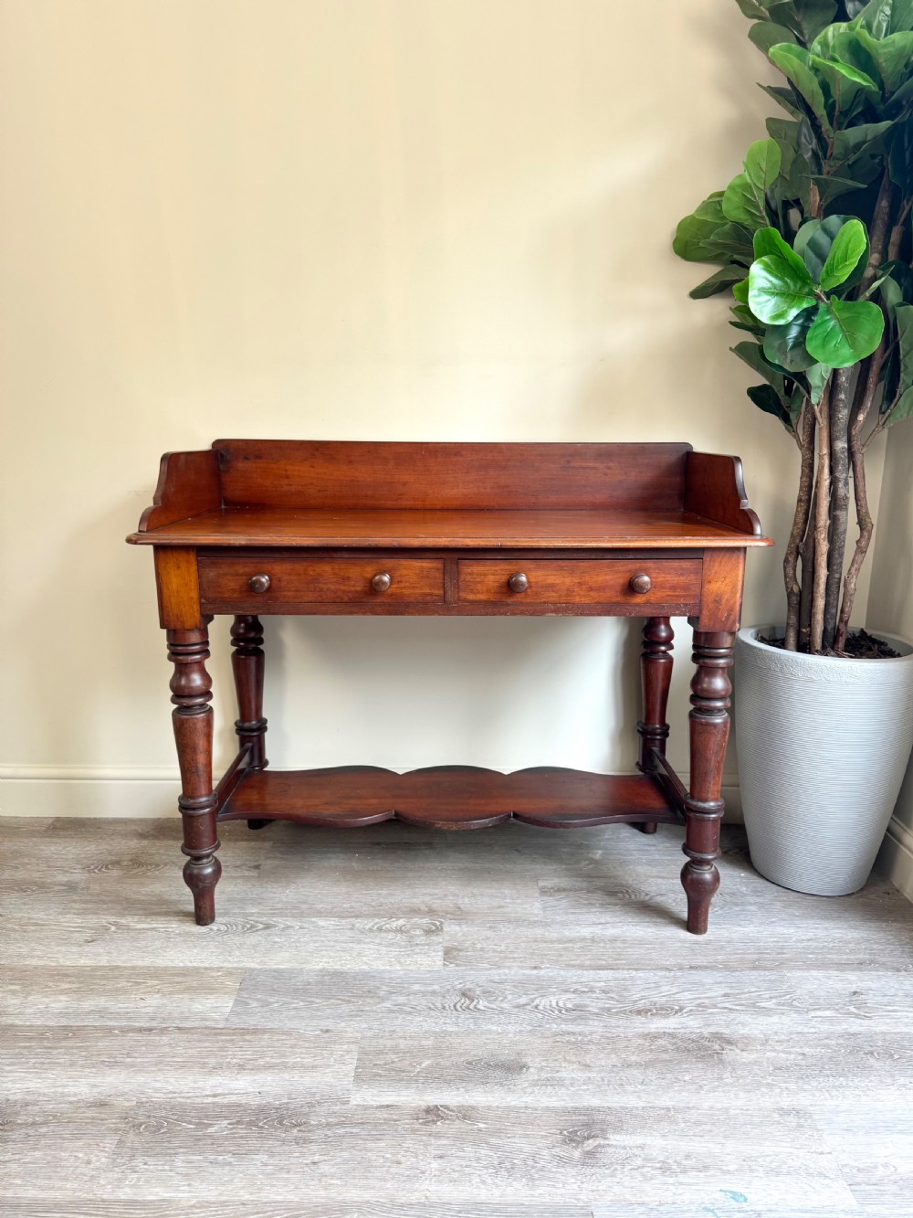 A warm brown mahogany console table featuring a rear splashback gallery, two drawers with wooden knobs, turned legs, and a shaped lower shelf. View V