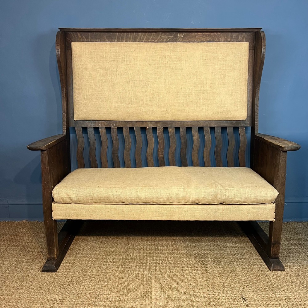 An antique Arts and Crafts dark oak high-back settle featuring vertical wavy slats and beige fabric upholstery.