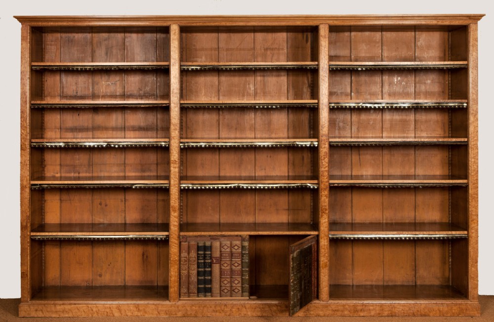 A large nineteenth-century open-front birdseye maple bookcase featuring three vertical sections, adjustable shelves with leather dust aprons, and a lower concealed compartment holding books.