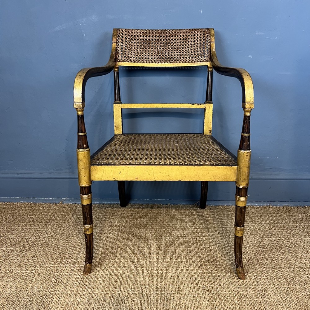 A Regency parcel gilt and faux rosewood armchair featuring a rectangular caned backrest, caned seat, sweeping arms, and turned legs with gold highlights.