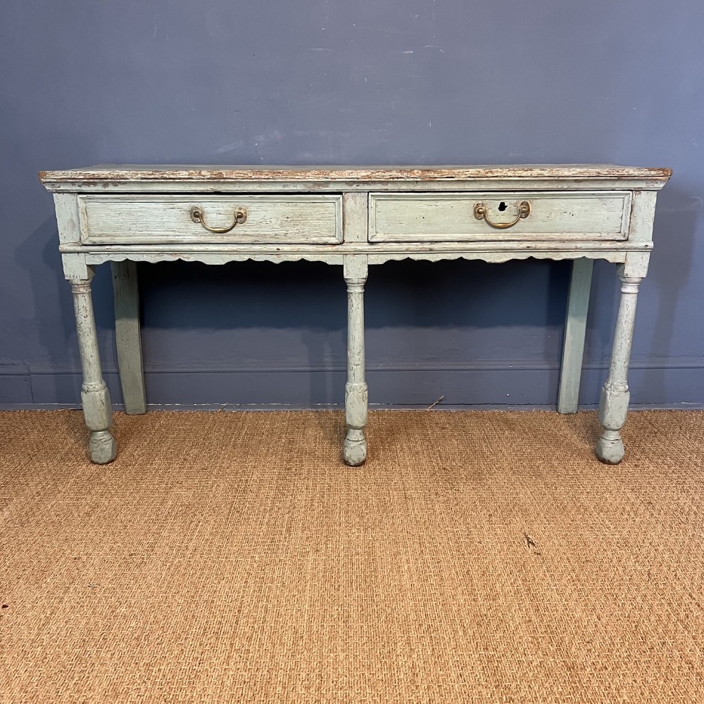 An early 18th-century painted pine dresser base featuring two wide drawers with brass handles, a scalloped apron, and turned front legs with a distressed pale blue-grey finish.