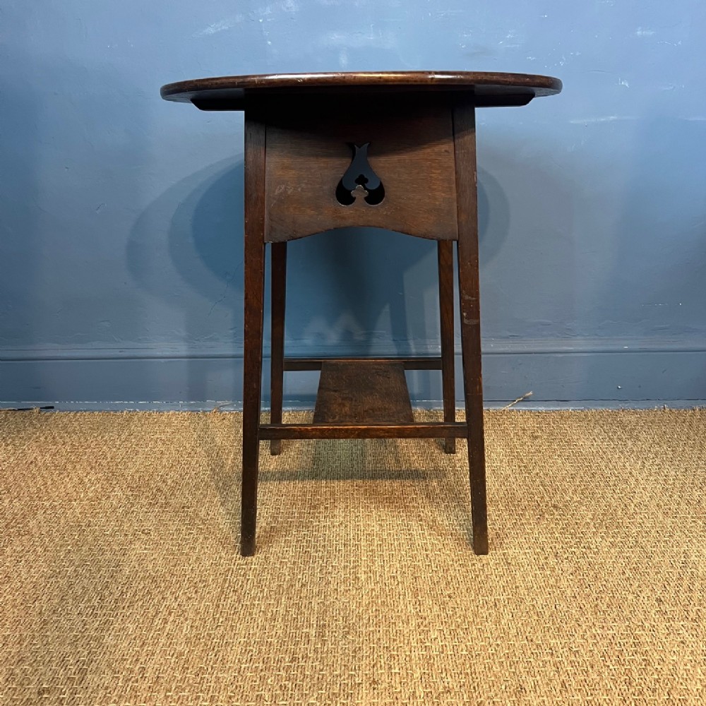 An antique dark oak circular Arts and Crafts two-tier side table featuring tapered legs and a pierced spade motif on the apron.