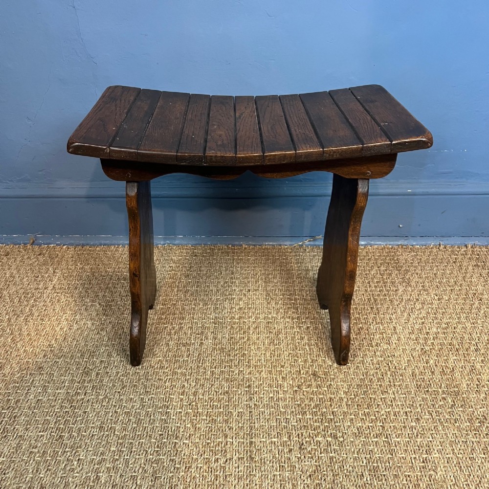 An Arts and Crafts dark oak stool featuring a concave slatted saddle seat supported by contoured side legs.