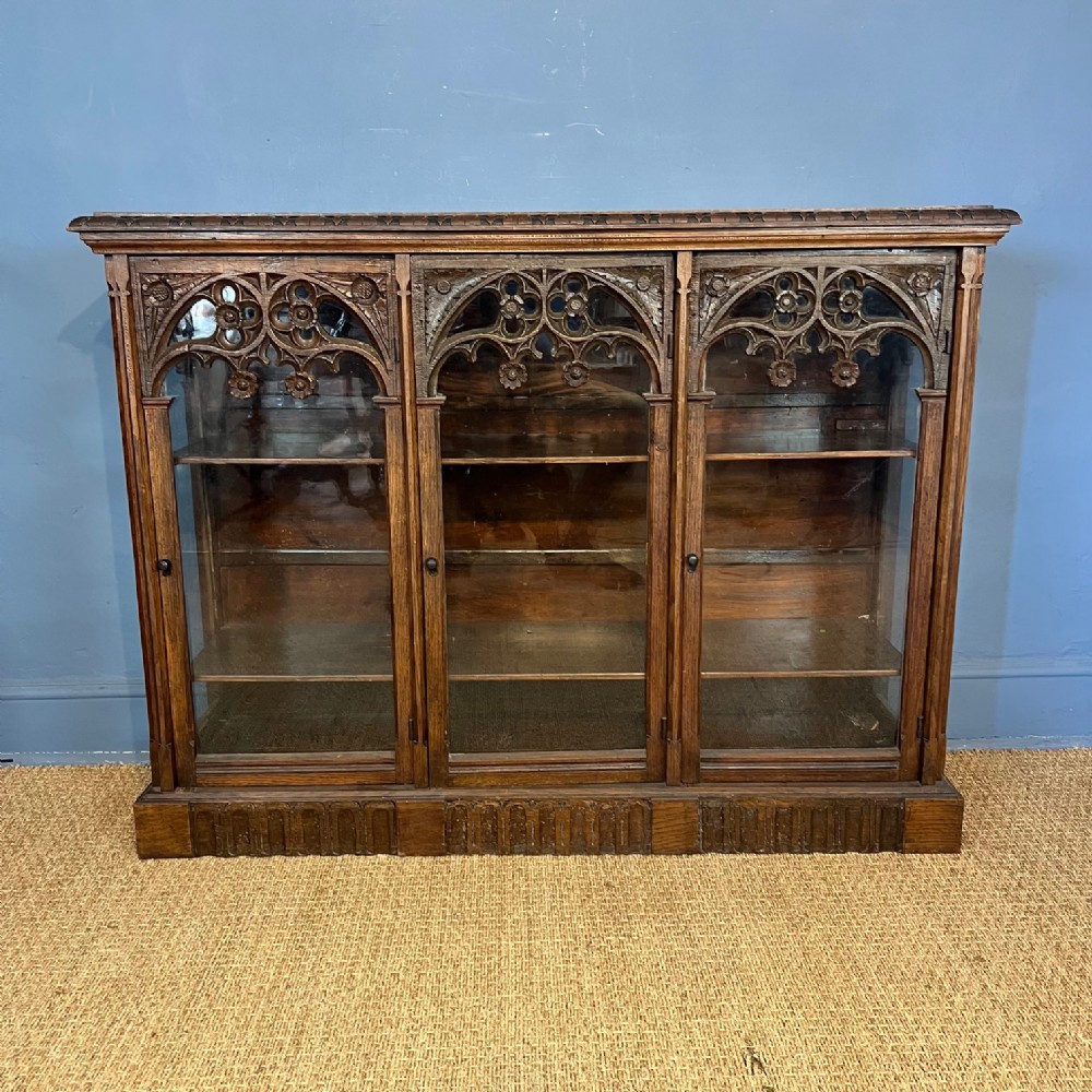 A mid-19th-century oak side cabinet with three glazed doors featuring carved Gothic tracery arches across the top.
