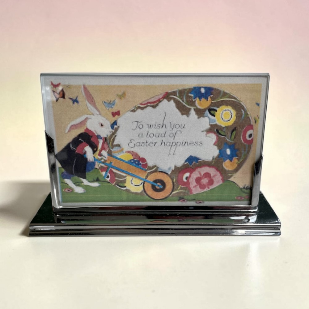 An Art Deco chrome freestanding photo frame featuring a stepped rectangular base holding a vintage postcard behind a glass panel.