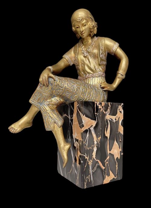 fig 1. A gilt bronze Art Deco sculpture of a seated female figure wearing patterned attire, perched atop a black and gold veined marble block.