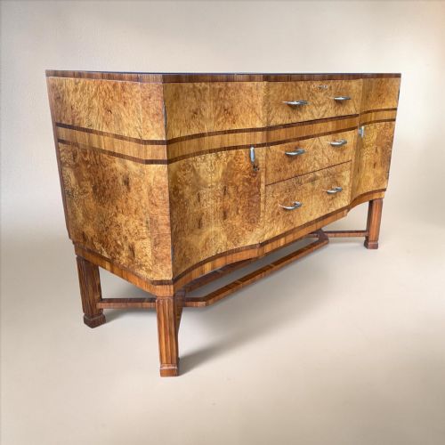 fig 1. An Art Deco burr walnut sideboard featuring a curved serpentine front, three central drawers with metal handles, side cupboards, and fluted legs joined by a stretcher.