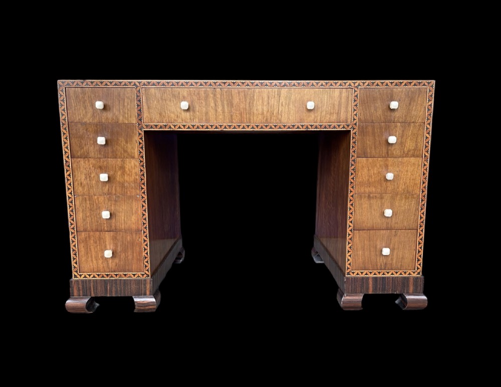 An Art Deco walnut kneehole desk featuring geometric inlaid borders, eleven drawers with square off-white knobs, and a plinth base with curved feet.