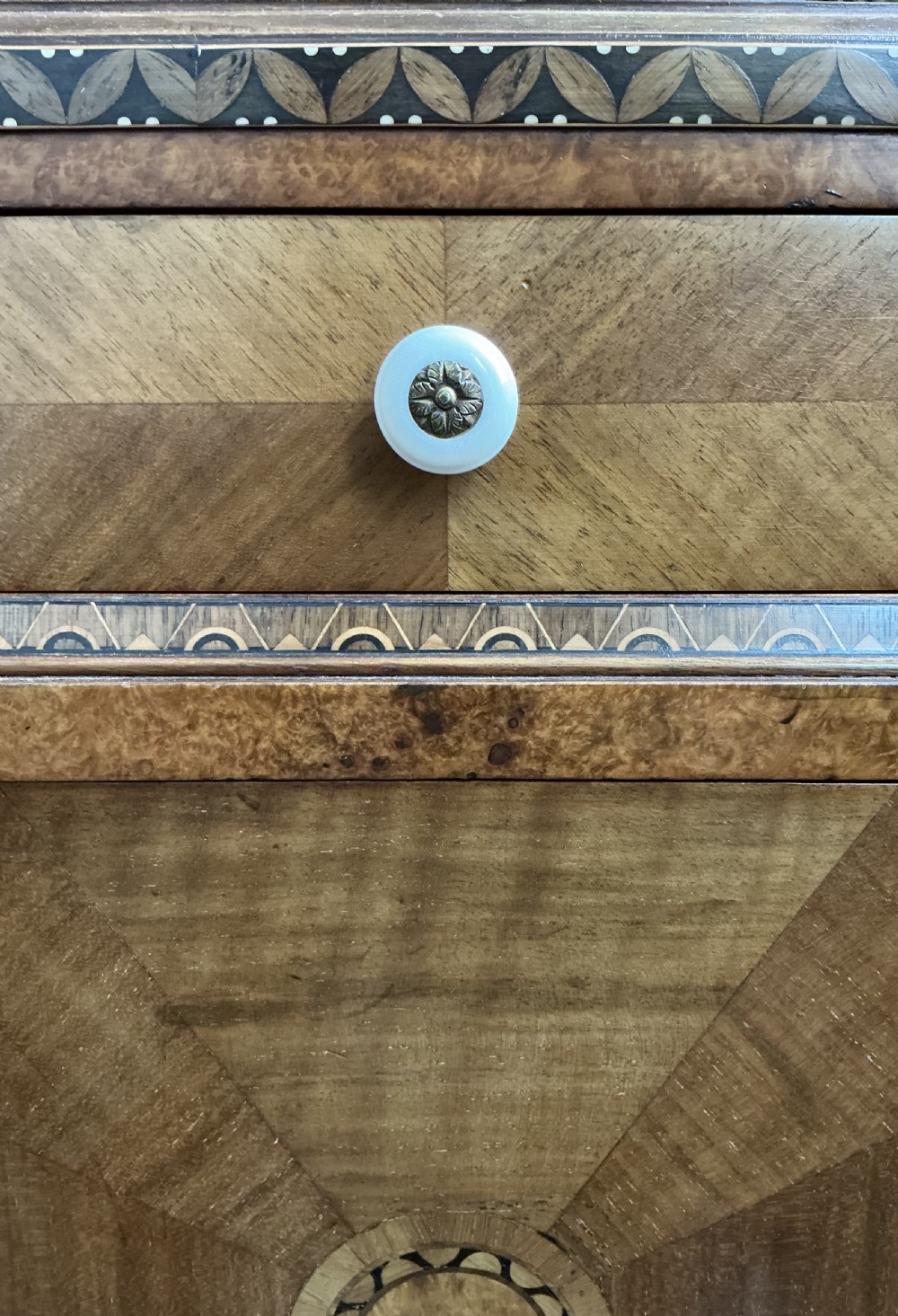 An Art Deco warm brown walnut bedside cabinet with geometric marquetry inlay, single top drawer, lower cupboard door, white ceramic knobs, and an X-stretcher base. View V