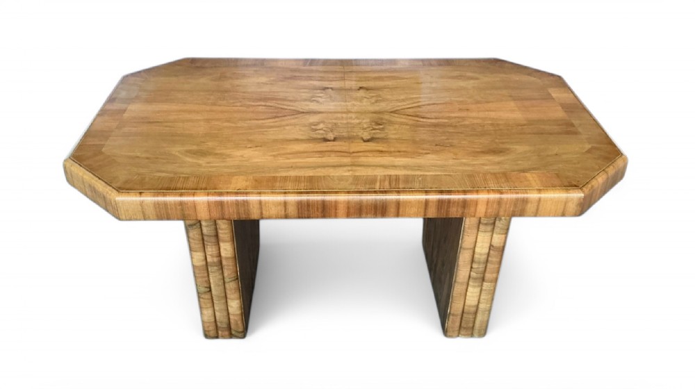 An Art Deco dining table featuring a figure-matched walnut top with canted corners and twin reeded pedestal bases.