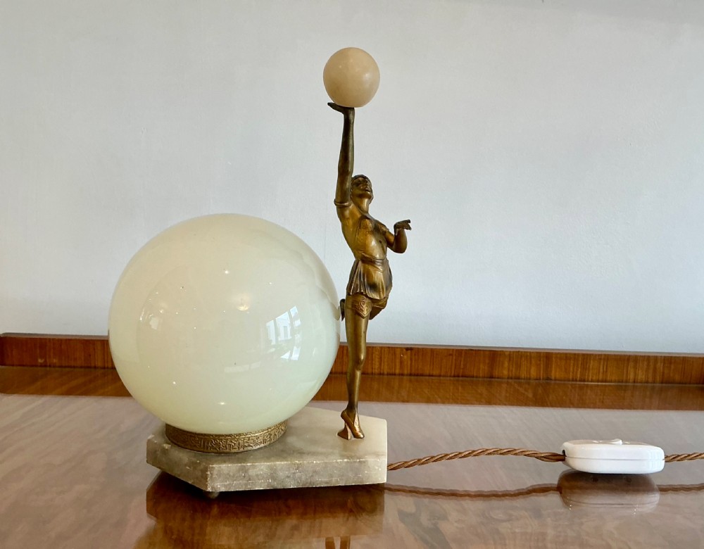 An Art Deco table lamp featuring a bronze figurine of a dancing lady posed beside a spherical frosted glass shade set on a marble base.