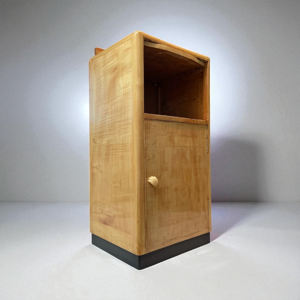 An Art Deco blonde wood bedside cabinet featuring an open upper compartment and a single cupboard door with a shell handle on a dark plinth base.