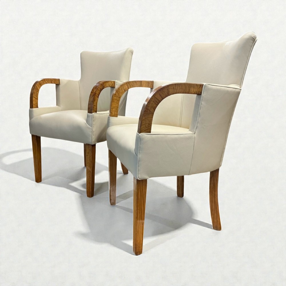 A pair of Art Deco armchairs upholstered in cream leather with curved walnut wooden arms and tapered legs.