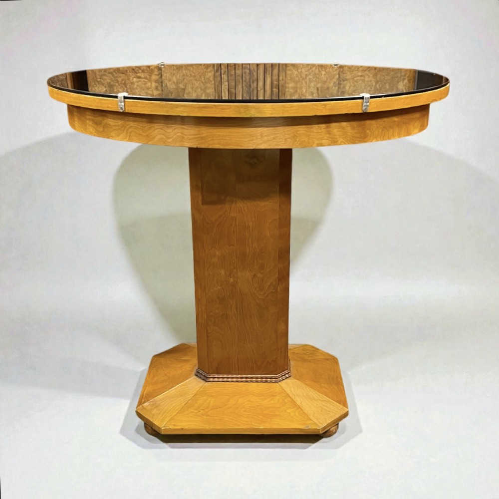 An Art Deco light wood centre table featuring a glass top secured by metal clips, resting on a central column and an octagonal base.