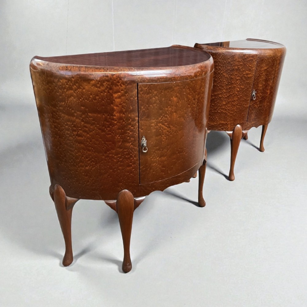 A pair of semi-circular Art Deco side cabinets featuring warm brown figured wood veneers, single curved cupboard doors, metal handles, and slender cabriole legs.