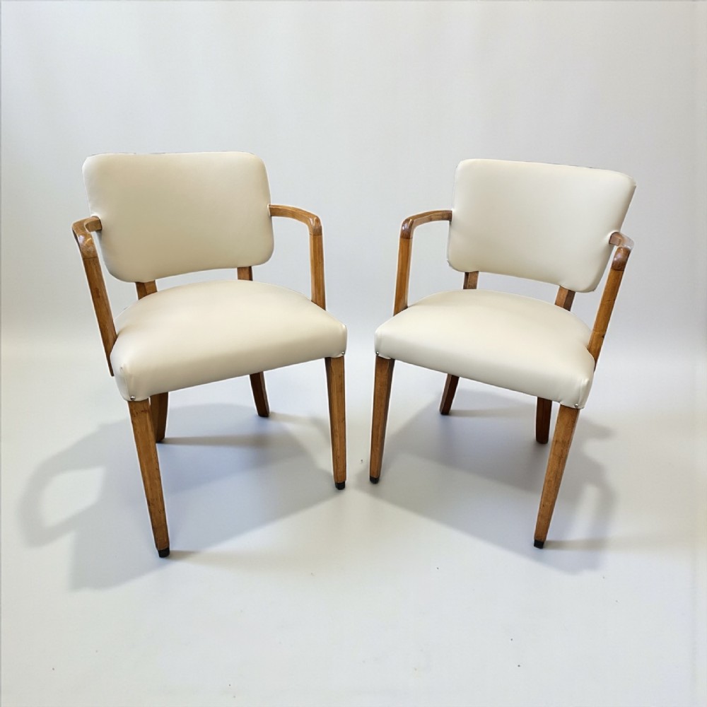 A pair of Art Deco armchairs featuring cream upholstery, curved wooden armrests, and tapered wooden legs against a plain background.