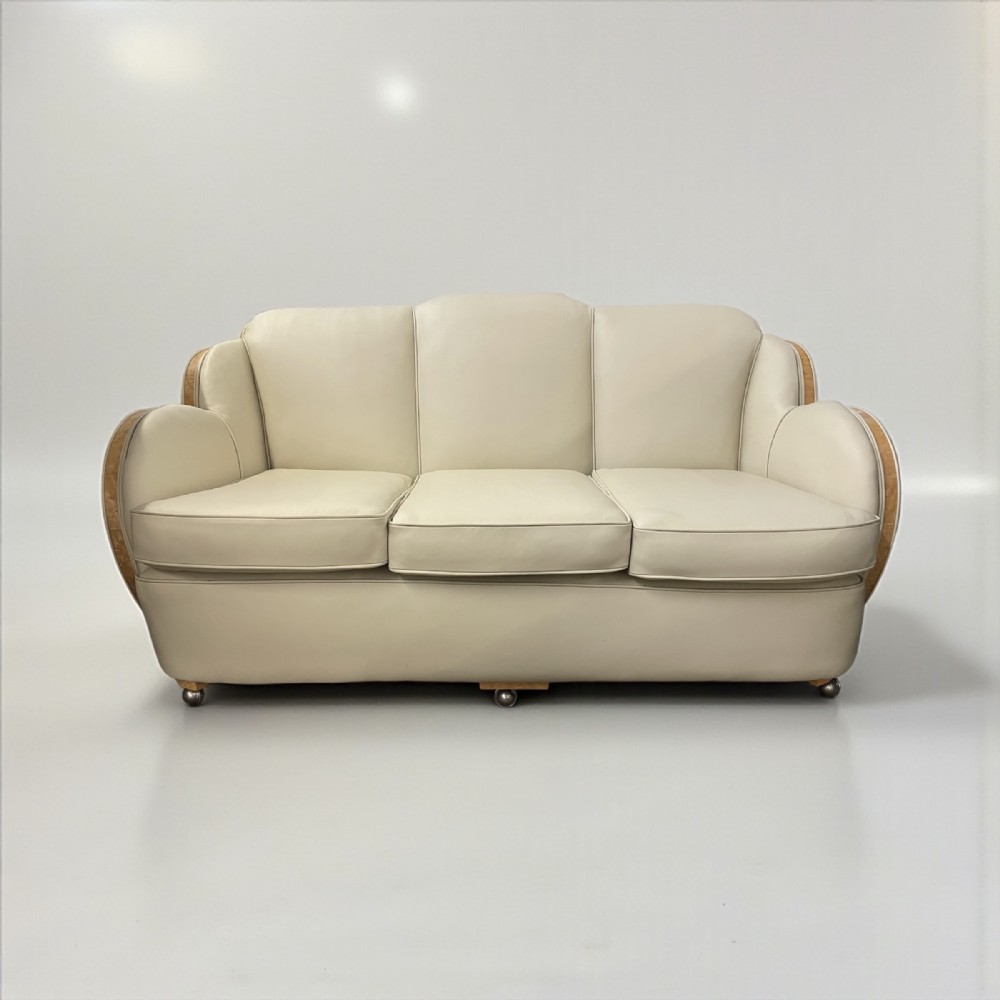 A three-seater Art Deco cloud sofa featuring cream leather upholstery and polished veneered timber arm supports.