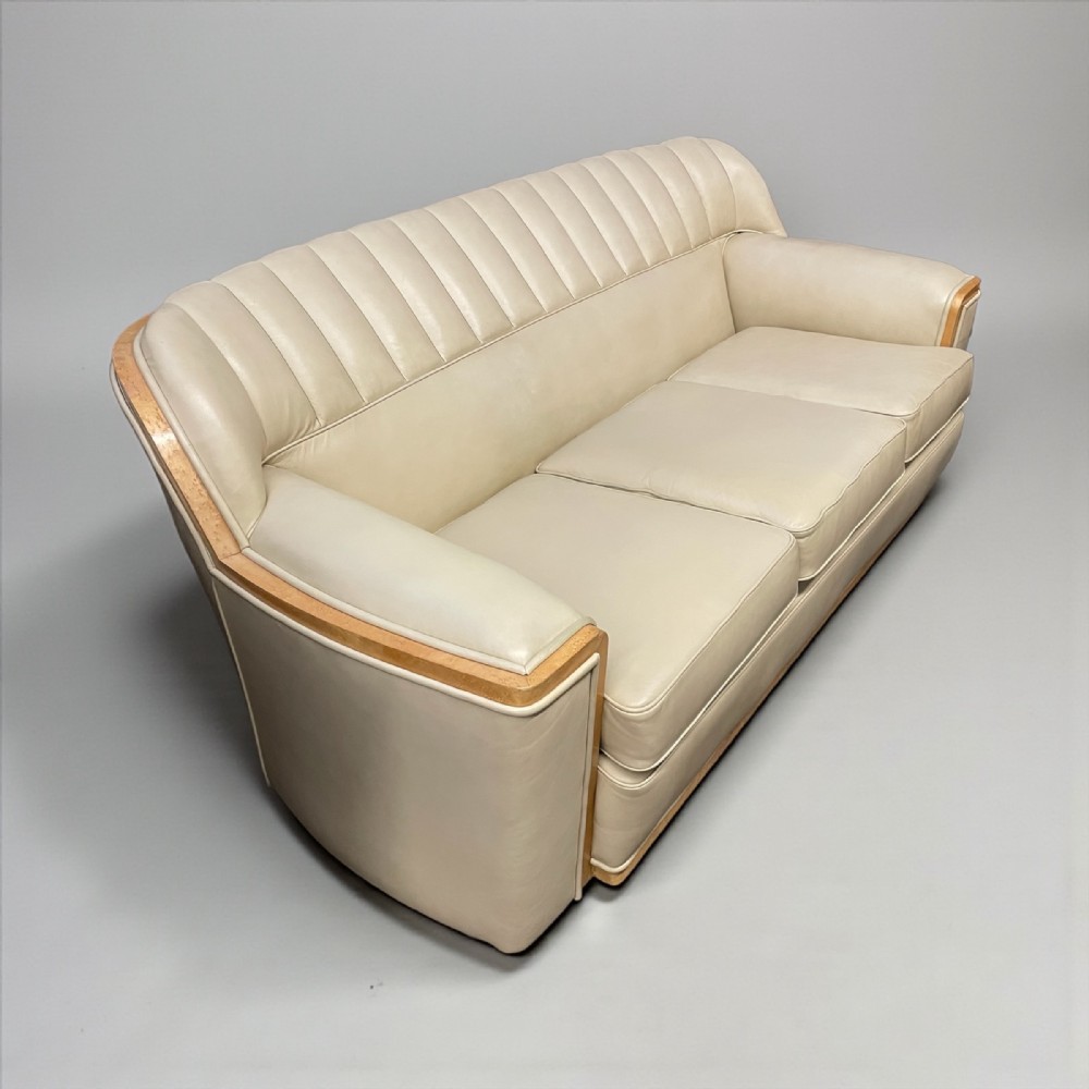 An Art Deco cream leather three-seater sofa featuring fluted back cushions and curved wood veneer arm trim.