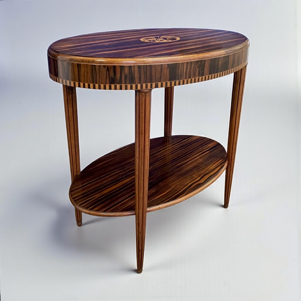 An oval Art Deco side table featuring dark striped wood veneer, a central floral inlay, a lower shelf tier, and slender fluted legs.