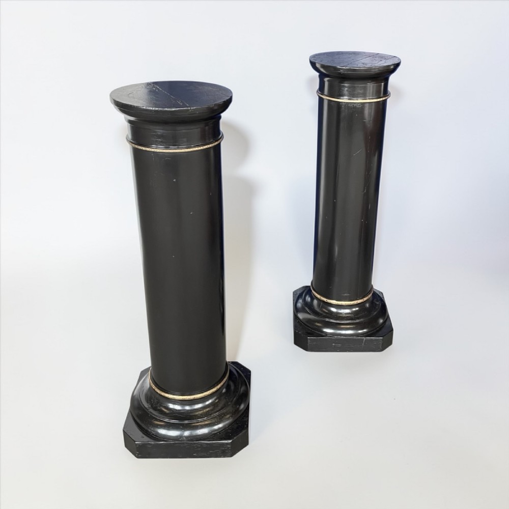 A pair of 19th-century ebonised wooden column pedestals featuring cylindrical shafts, stepped octagonal bases, circular tops, and narrow gilt ring detailing.