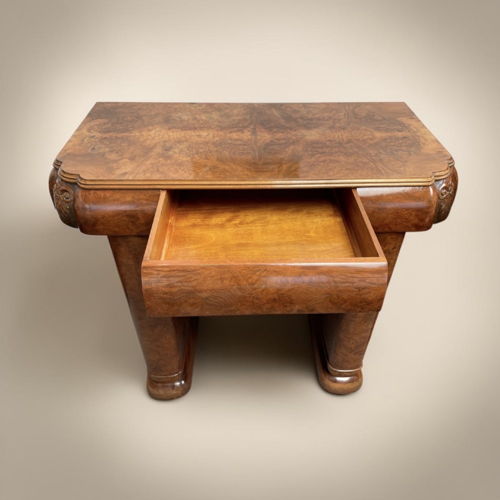 An Art Deco burr walnut console table featuring a single open drawer, streamlined curved shoulders, and thick tapered legs with carved detailing.