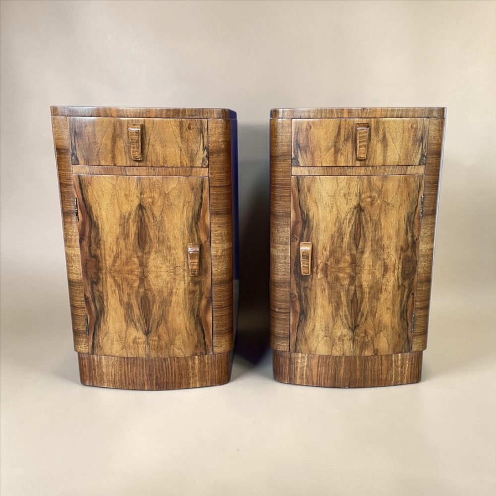 A matching pair of Art Deco walnut bedside cabinets featuring book-matched wood grain, single top drawers, rounded sides, and lower cupboard doors.