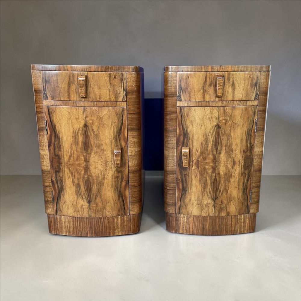 A matching pair of Art Deco walnut bedside cabinets featuring book-matched wood grain, single top drawers, rounded sides, and lower cupboard doors. View X