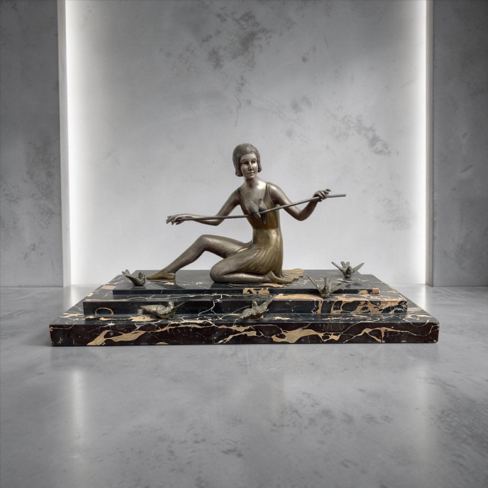 An Art Deco bronze sculpture depicting a seated woman holding a thin rod surrounded by small birds on a tiered black and gold marble base.