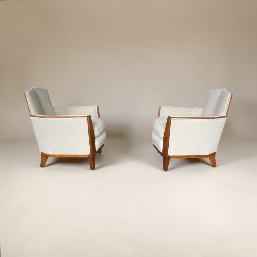 A pair of Art Deco lounge armchairs with light cream fabric upholstery and geometric dark wood arm trims.