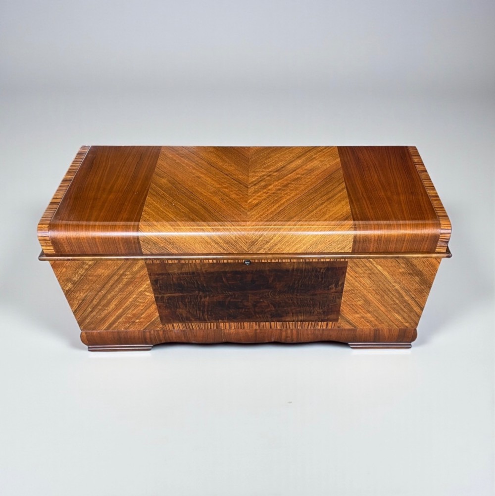 An Art Deco timber blanket chest featuring geometric chevron wood veneers and rounded waterfall edges on a stepped plinth base. View III