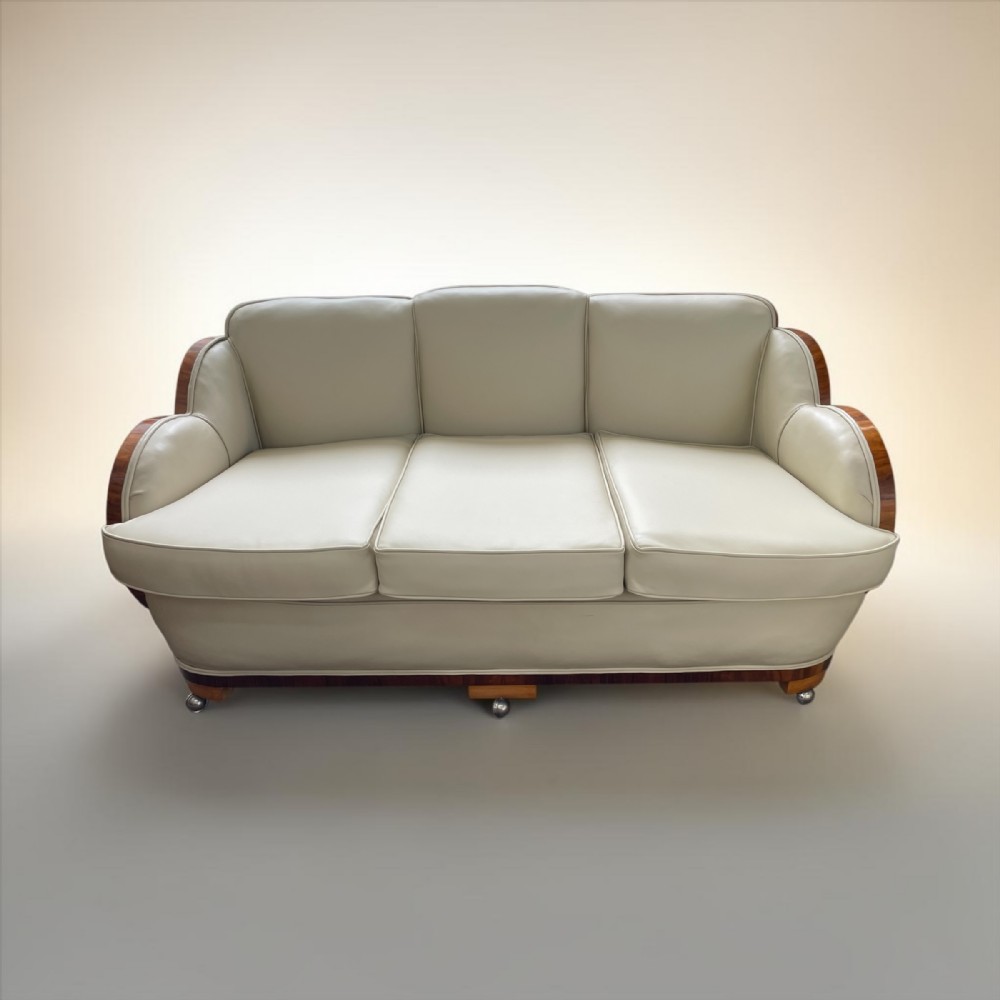 An Art Deco three-seater sofa upholstered in cream leather with curved walnut-veneered arms and a walnut base trim.