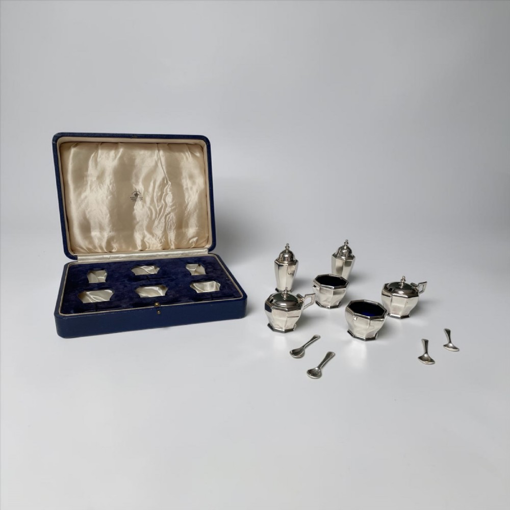 A complete Art Deco sterling silver condiment set including faceted pepper casters, salt cellars with cobalt blue glass liners, mustard pots, and spoons alongside a fitted blue presentation box.