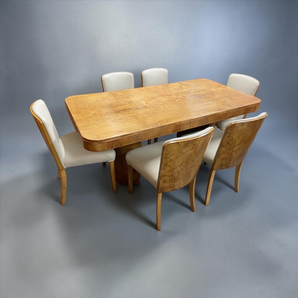 An Art Deco burr walnut dining table featuring rounded corners and a central pedestal base, accompanied by six matching cloud-back chairs upholstered in off-white leather.