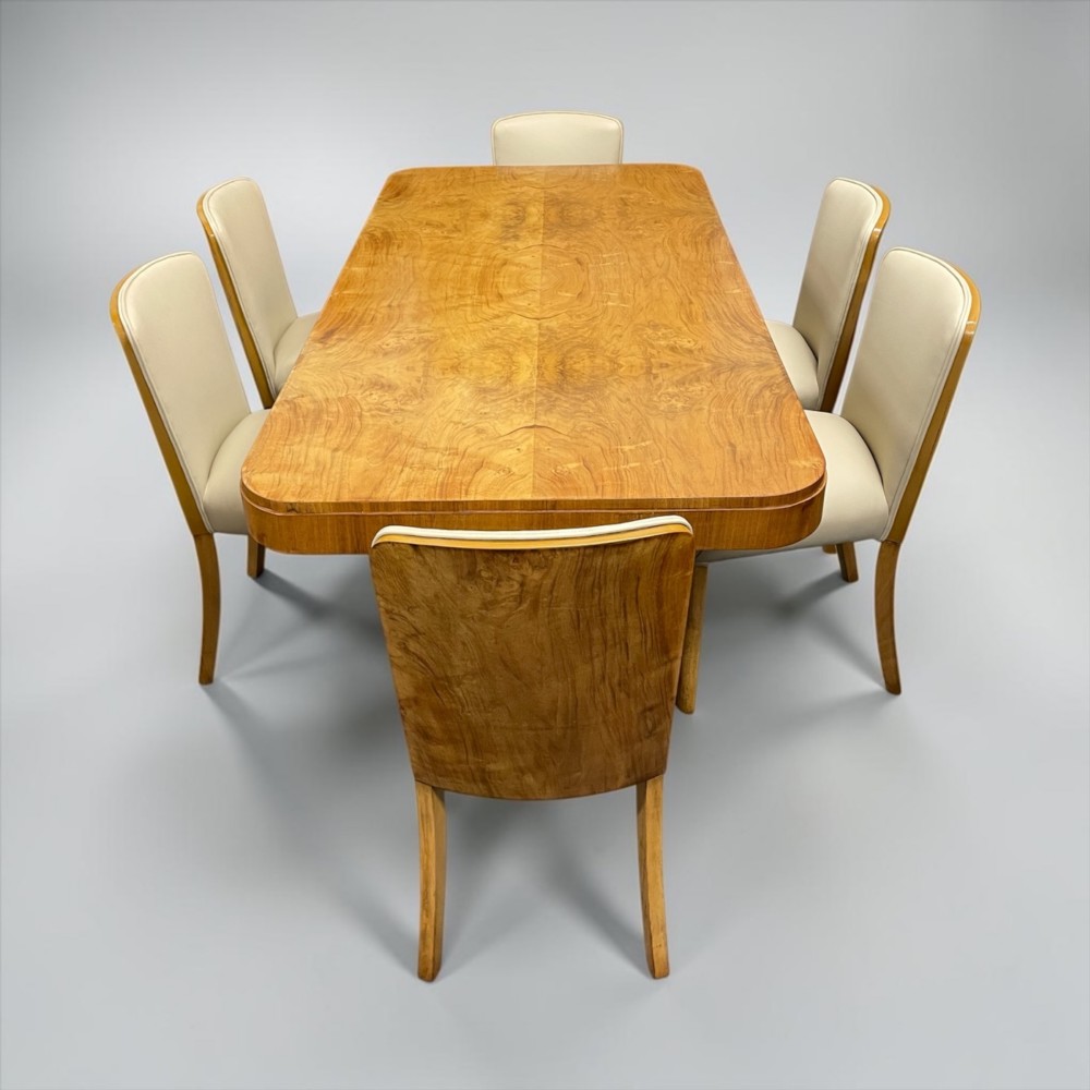 An Art Deco burr walnut dining table featuring rounded corners and a central pedestal base, accompanied by six matching cloud-back chairs upholstered in off-white leather. View XV