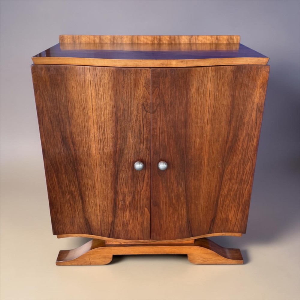 An Art Deco walnut sheet music cabinet with a curved front profile, double doors, metal knobs, and a curved base.