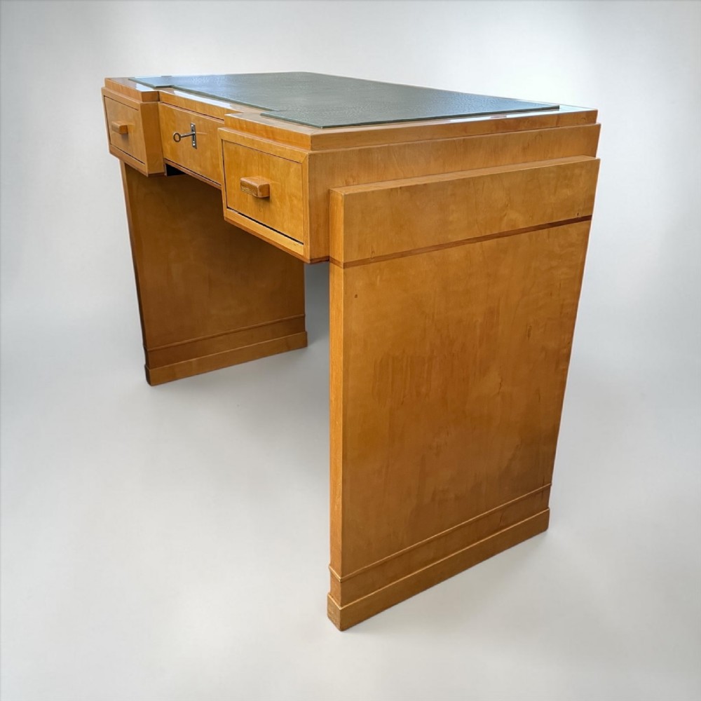 An Art Deco light wood writing desk featuring an inset leather writing surface, three drawers with rectangular wooden pulls, and a central keyhole.
