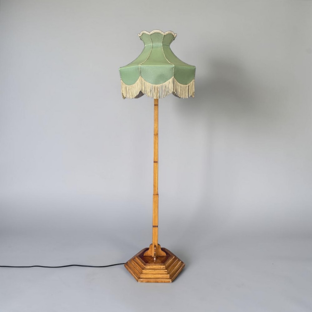 An Art Deco wooden standard lamp featuring a stepped octagonal base, a tall central stem with geometric inlay, and a green scalloped fabric shade with cream tassel fringe.
