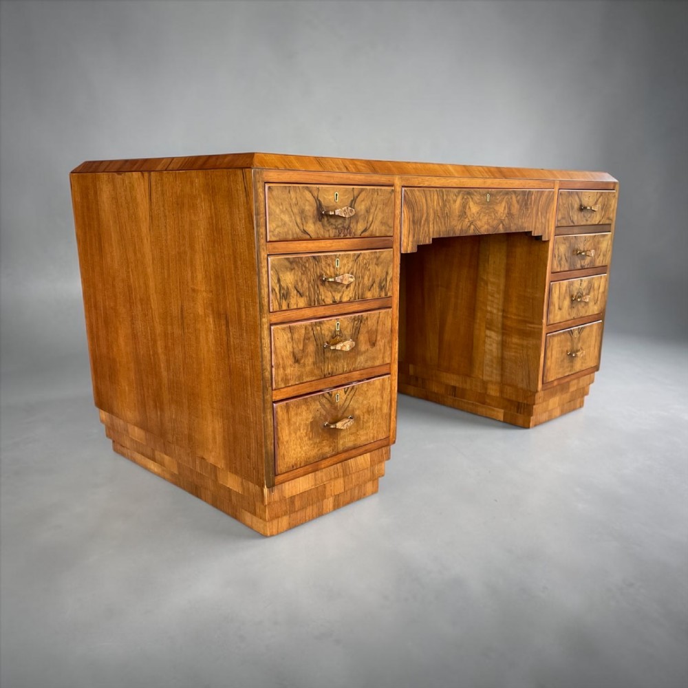 An Art Deco walnut pedestal desk featuring a stepped plinth base, book-matched veneer drawers, and a stepped central kneehole arch.