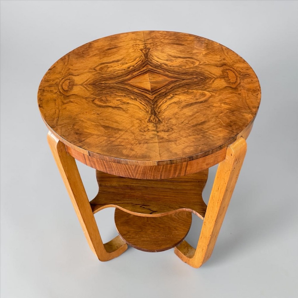 An Art Deco circular walnut occasional table featuring a quarter-booked grain veneer top, bent legs, and lower tiered shelving.