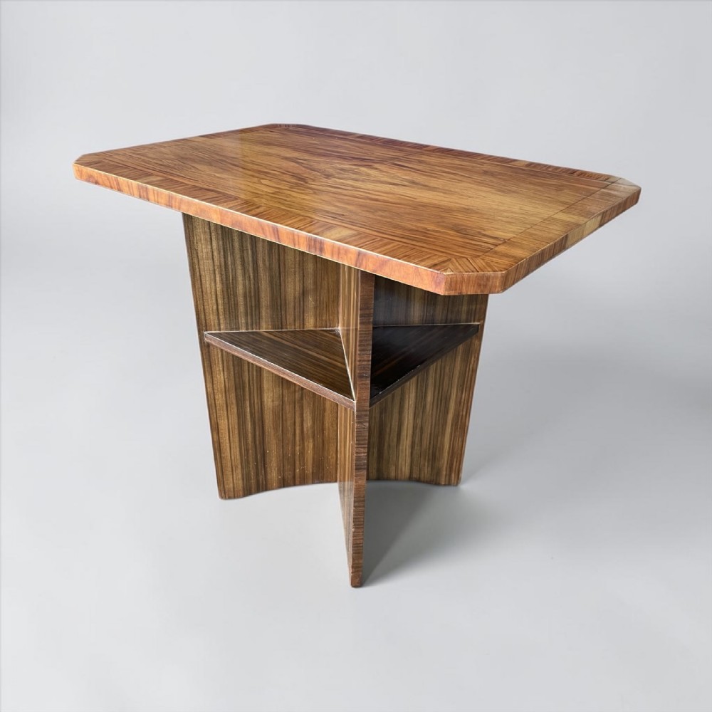 An Art Deco coffee table with a polished figured wood veneer surface, canted corners, a geometric cross-shaped base, and a triangular lower shelf.