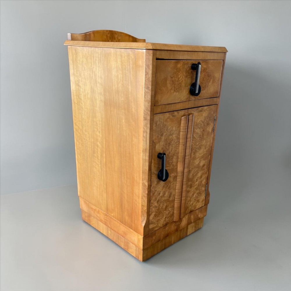 An Art Deco blonde burr wood bedside cabinet featuring a top drawer, lower cupboard door with curved detailing, and vertical black handles.