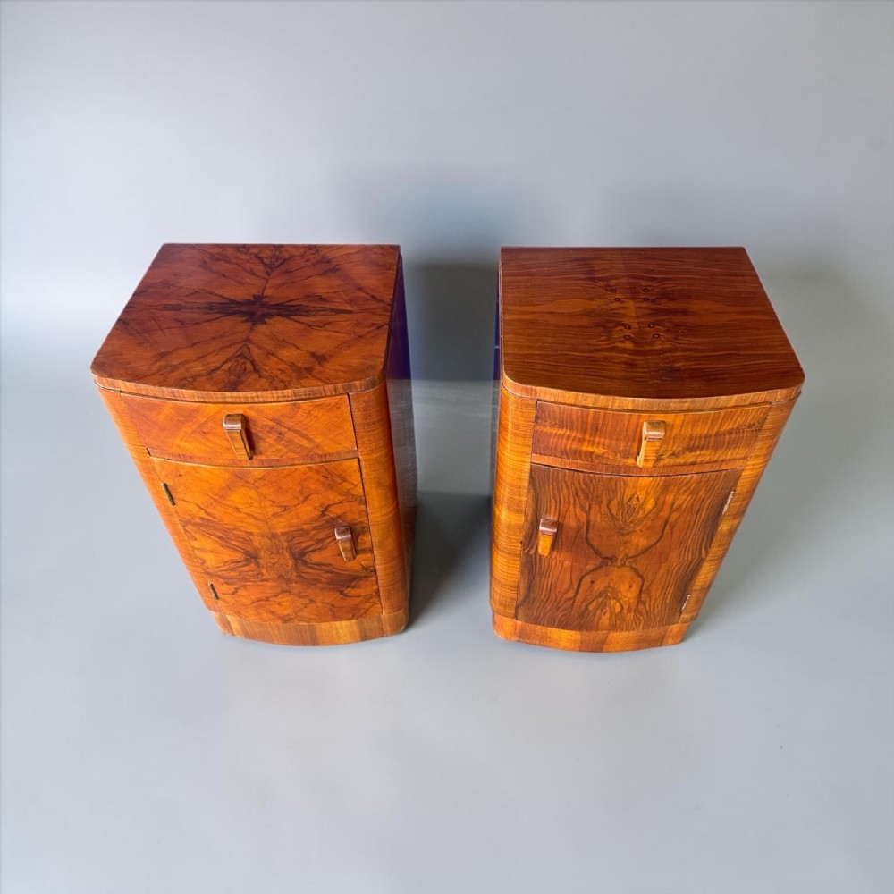 A pair of Art Deco walnut bedside tables featuring curved bow fronts, single top drawers, lower cupboard doors, and bookmatched timber veneers.