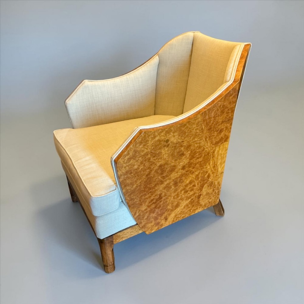 An Art Deco style armchair with angled burr wood side panels and cream textile upholstery.