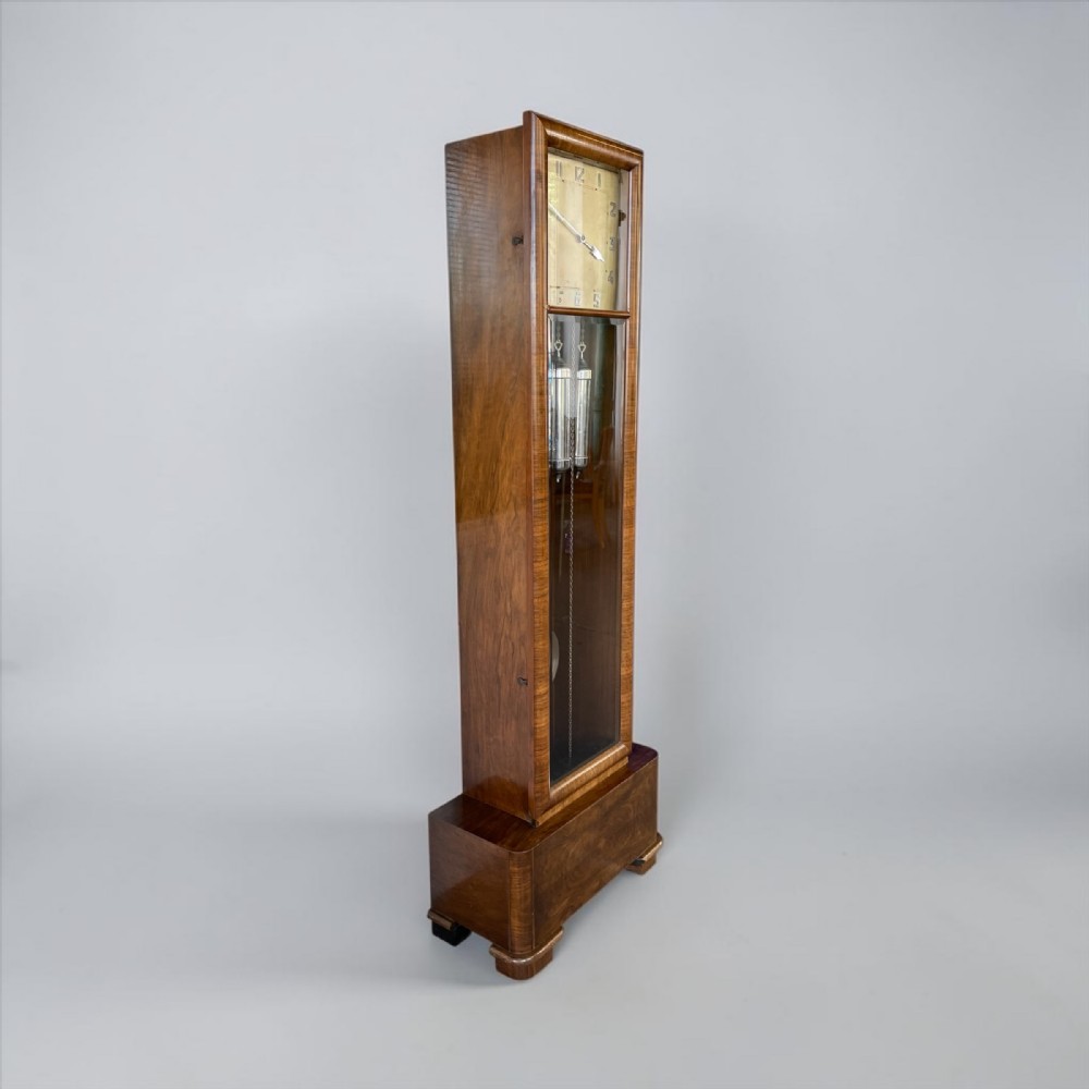 An Art Deco walnut longcase clock featuring a rectangular glass door, visible chrome weights, a square silvered dial, and a stepped plinth base.